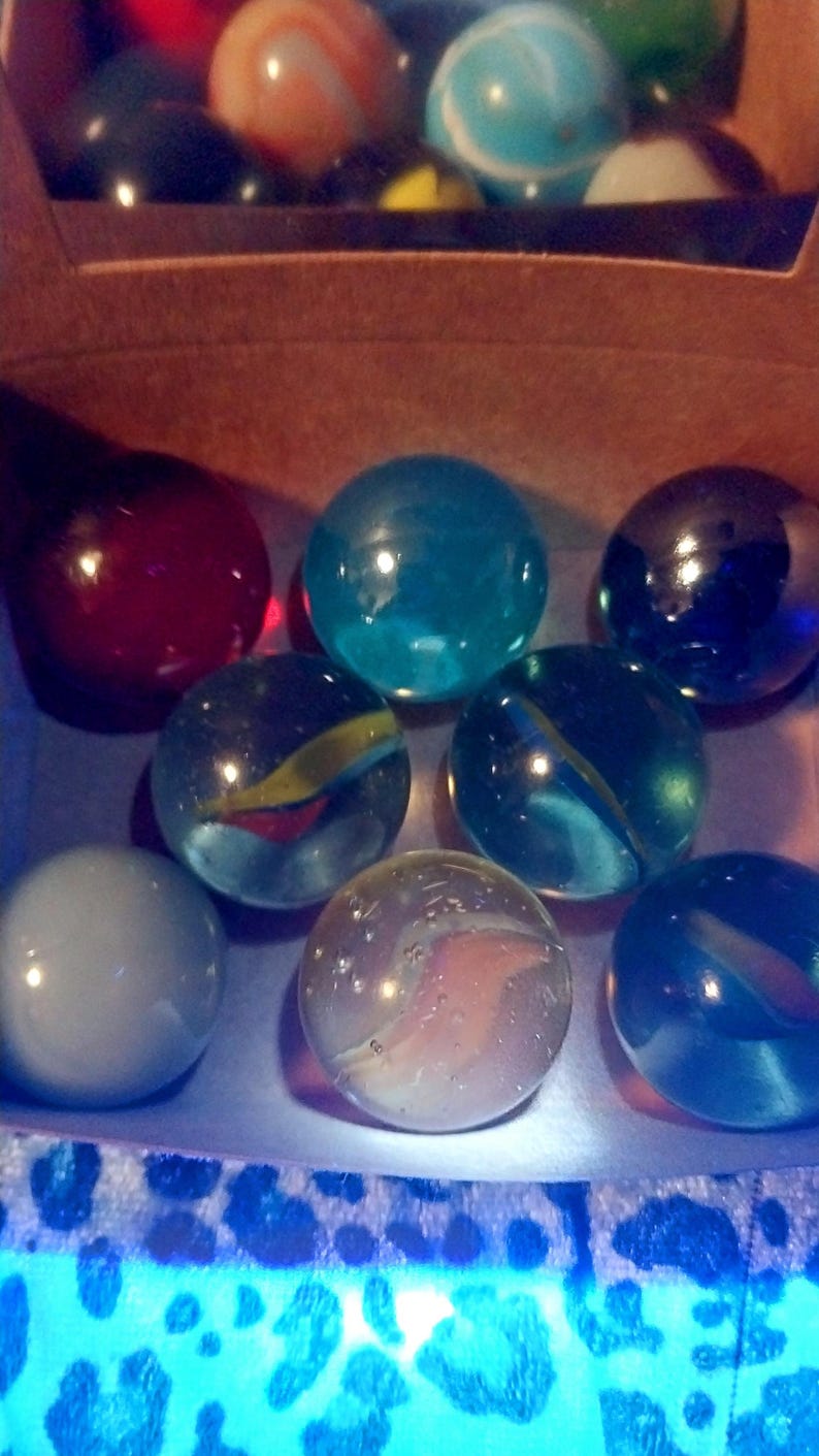 May include: A collection of colorful glass marbles in various shades of blue, red, and clear. Some marbles have internal swirls of color. The marbles are in a cardboard box with a patterned edge.