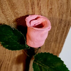 May include: A pink ceramic rose with green leaves attached to a wooden surface.