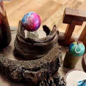 May include: A collection of handcrafted decorative items, including painted wooden spheres in shades of blue, pink, and green. The spheres are displayed on a wooden surface with natural wood elements, such as bark and a pine cone, creating a rustic aesthetic.