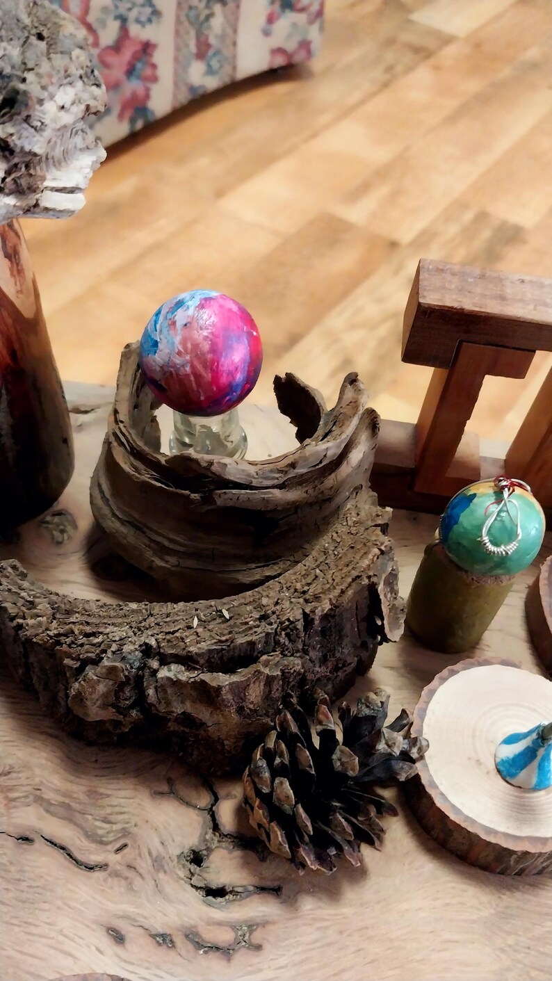 May include: A collection of handcrafted decorative items, including painted wooden spheres in shades of blue, pink, and green. The spheres are displayed on a wooden surface with natural wood elements, such as bark and a pine cone, creating a rustic aesthetic.