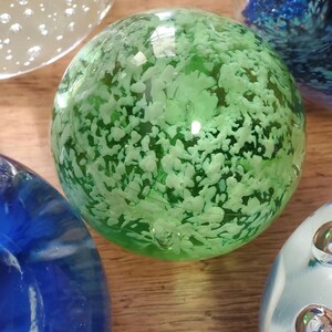 May include: A selection of decorative glass spheres. One sphere is a vibrant green with white inclusions. Other spheres are blue and clear, with bubble and swirl designs. The spheres are on a wooden surface.