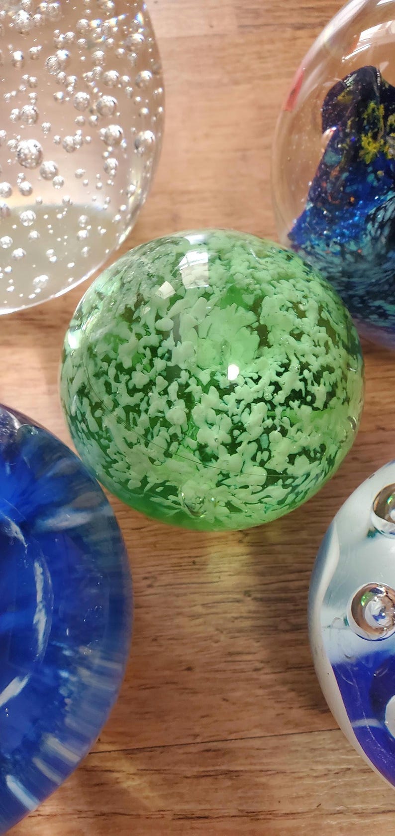 May include: A selection of decorative glass spheres. One sphere is a vibrant green with white inclusions. Other spheres are blue and clear, with bubble and swirl designs. The spheres are on a wooden surface.