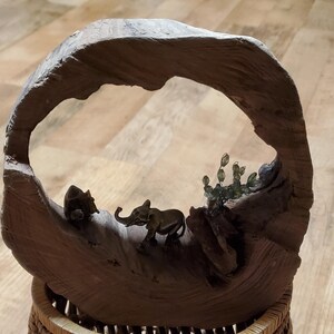 May include: A decorative wooden sculpture featuring a carved arch with miniature bronze elephant and rhino figurines. The sculpture includes a small cactus and sits atop a woven basket base. The wood has a natural, weathered appearance.