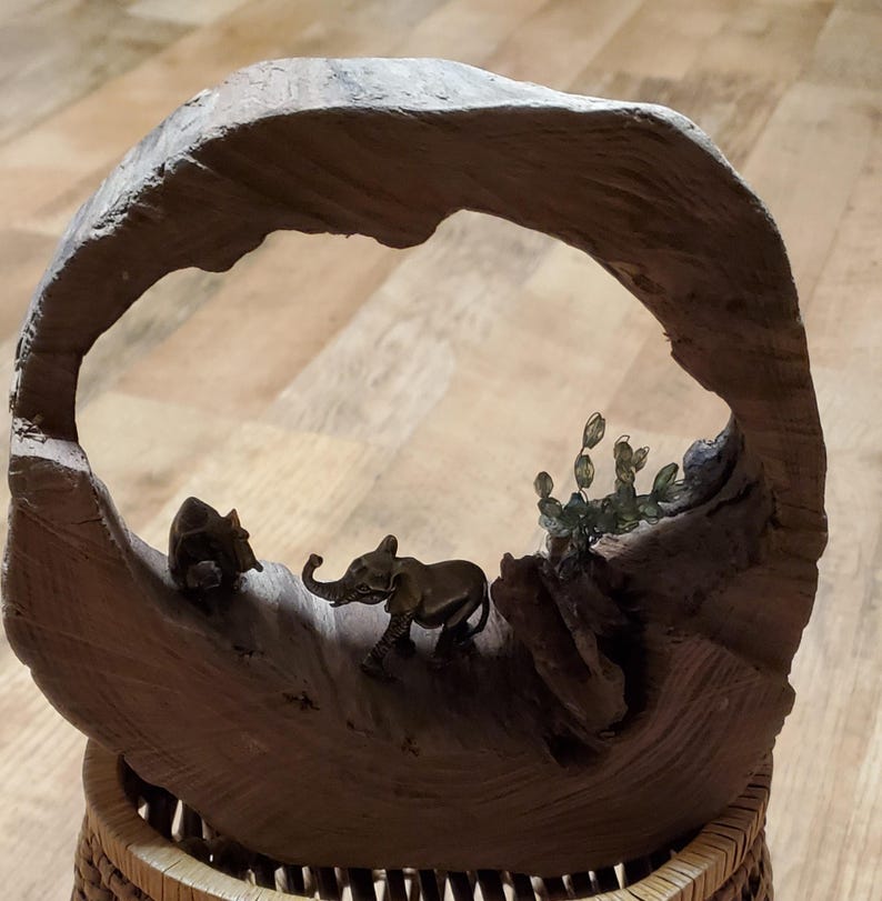 May include: A decorative wooden sculpture featuring a carved arch with miniature bronze elephant and rhino figurines. The sculpture includes a small cactus and sits atop a woven basket base. The wood has a natural, weathered appearance.