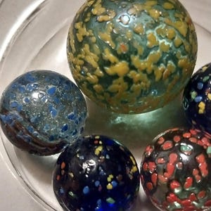 Op de afbeelding: Vijf glazen knikkers met verschillende patronen en kleuren. De knikkers zijn blauw, groen, rood, zwart en wit met spikkels.