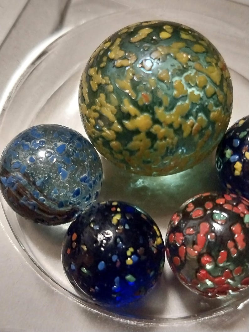 Op de afbeelding: Vijf glazen knikkers met verschillende patronen en kleuren. De knikkers zijn blauw, groen, rood, zwart en wit met spikkels.