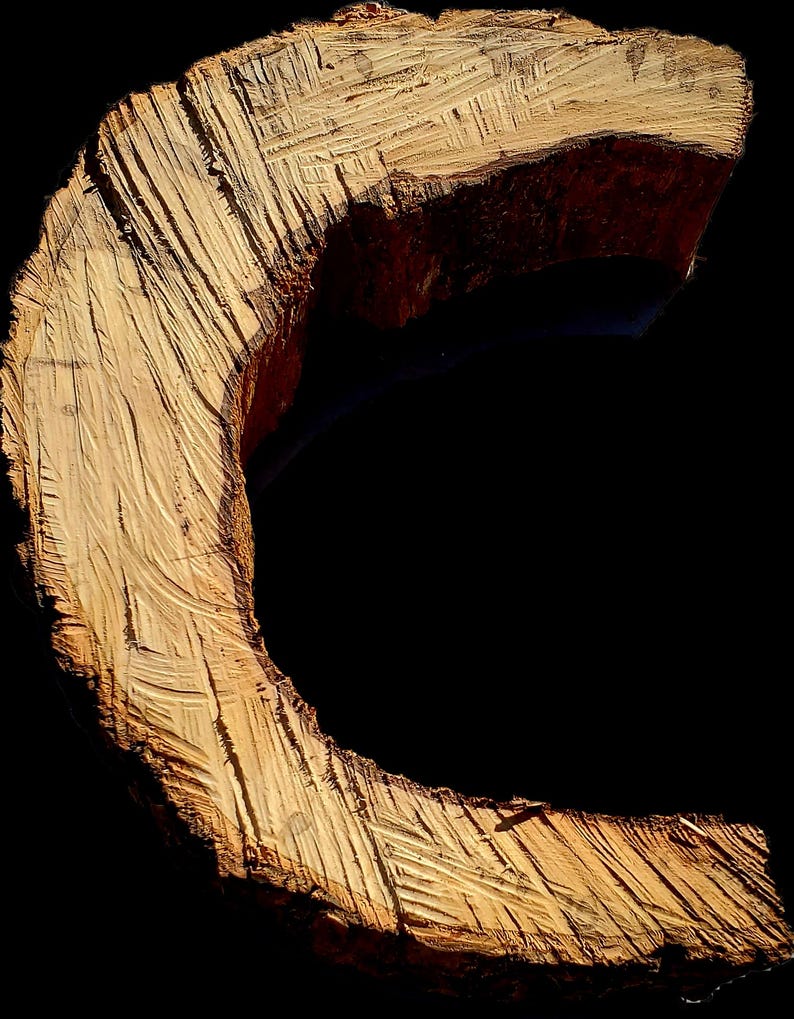 K&ouml;nnte beinhalten: Eine Nahaufnahme eines Holzstammes, der zu einem C geformt wurde. Das Holz ist hellbraun und hat eine raue Textur. Der Stamm ist vor einem schwarzen Hintergrund platziert.