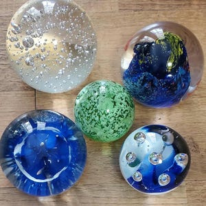 May include: Five decorative glass paperweights. One is clear with bubbles, another has blue and yellow swirls, a third is green with a speckled pattern, and the other two are blue and white with decorative elements.