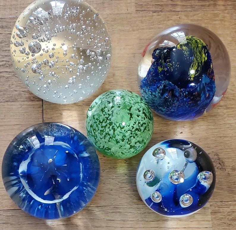 May include: Five decorative glass paperweights. One is clear with bubbles, another has blue and yellow swirls, a third is green with a speckled pattern, and the other two are blue and white with decorative elements.