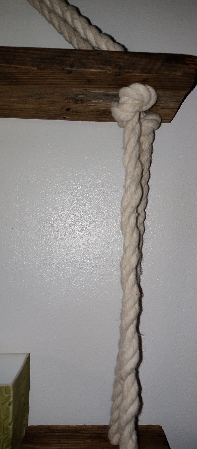 May include: A white rope is tied to a wooden shelf. The rope is knotted and hangs down from the shelf.