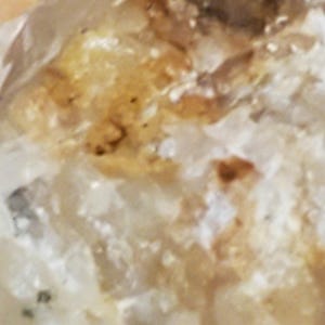 May include: Close-up of a raw, natural quartz crystal. The crystal has a translucent, milky white base with areas of light brown and amber coloration. The surface has a rough, textured appearance.