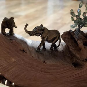 May include: A decorative wooden sculpture featuring two small bronze elephant figurines. One elephant stands upright, while the other has its trunk raised. A small, beaded tree is also present, adding to the natural theme.