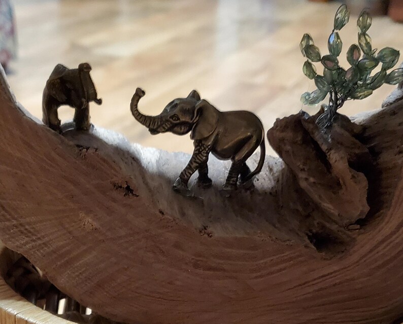 May include: A decorative wooden sculpture featuring two small bronze elephant figurines. One elephant stands upright, while the other has its trunk raised. A small, beaded tree is also present, adding to the natural theme.