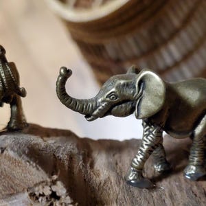 May include: Two small bronze elephant figurines on a piece of driftwood. One elephant is standing, and the other is kneeling. The elephants have detailed trunks, ears, and legs. The background is blurred, with a woven basket and a small green plant visible.