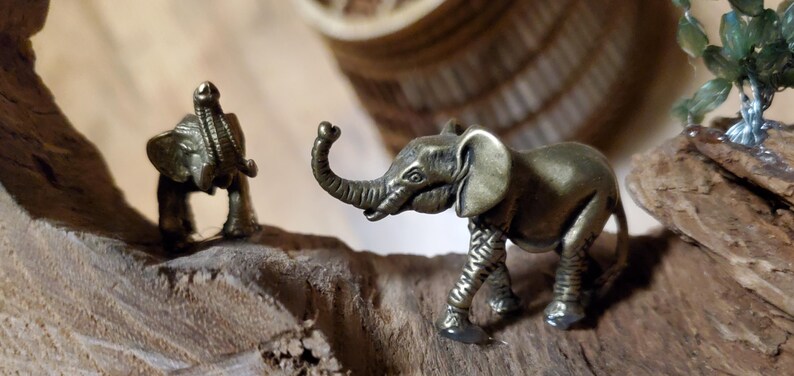 May include: Two small bronze elephant figurines on a piece of driftwood. One elephant is standing, and the other is kneeling. The elephants have detailed trunks, ears, and legs. The background is blurred, with a woven basket and a small green plant visible.