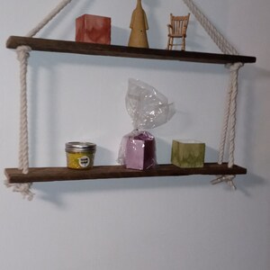 May include: Two wooden shelves with rope hangers, featuring a small wooden chair, a wooden figurine, a jar of yellow candle, a purple box, and a green bar of soap.
