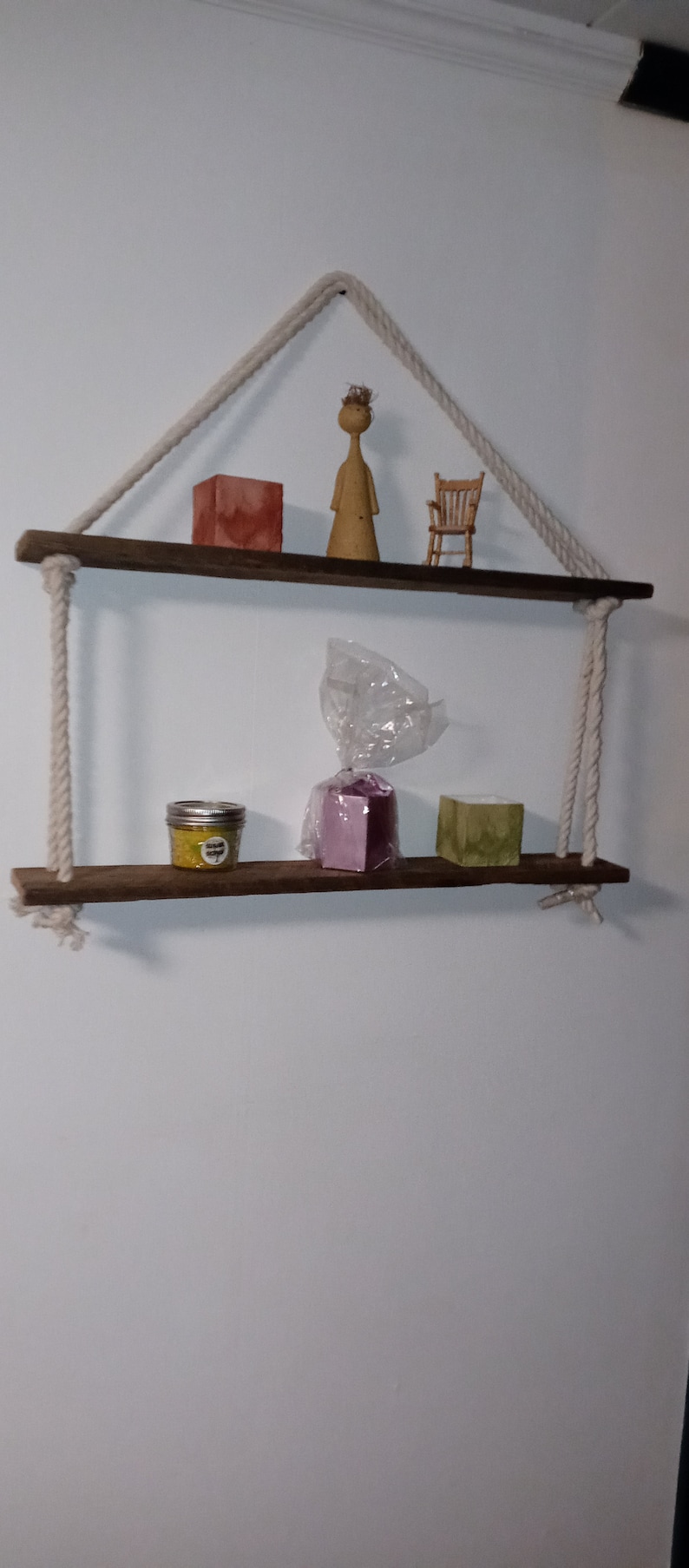 May include: Two wooden shelves with rope hangers, featuring a small wooden chair, a wooden figurine, a jar of yellow candle, a purple box, and a green bar of soap.