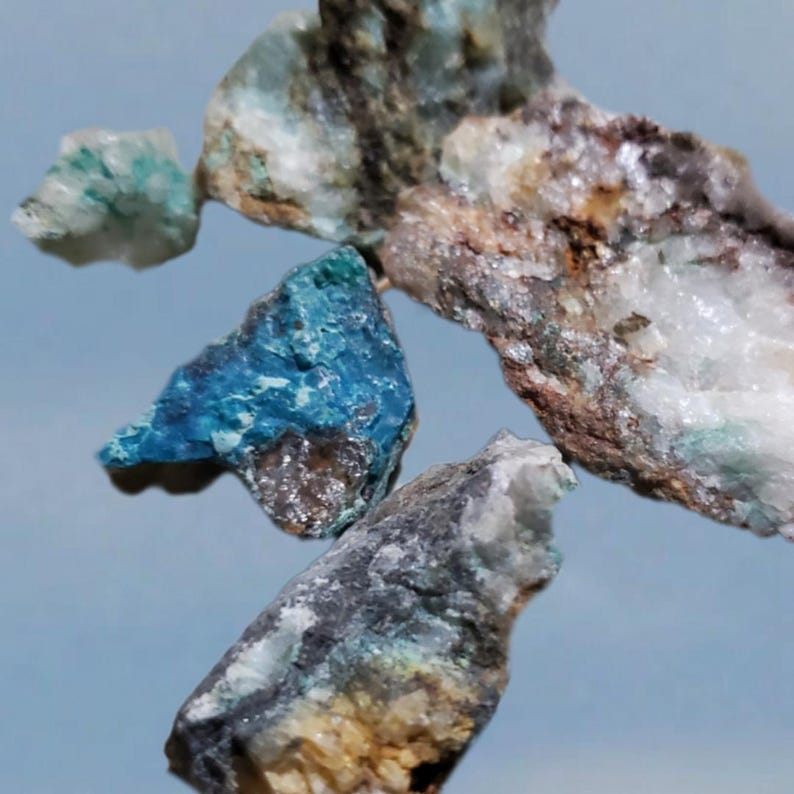 May include: A collection of rough, natural mineral specimens. The rocks are a mix of blue, green, white, and brown. The rocks are small and appear to be tumbled.
