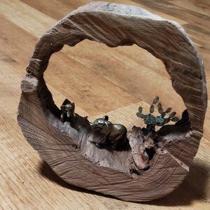 May include: A wooden sculpture featuring a circular frame with a carved-out interior. Inside, there are small brass elephant figurines and a metal cactus. The wood has a natural, light brown color.