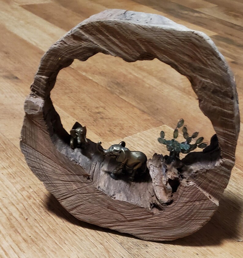 May include: A wooden sculpture featuring a circular frame with a carved-out interior. Inside, there are small brass elephant figurines and a metal cactus. The wood has a natural, light brown color.
