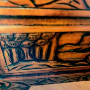 May include: Close-up of a wooden furniture piece with intricate, dark-colored carvings. The wood has a warm, orange-brown hue, and the carvings feature detailed patterns and shapes, creating a textured surface.