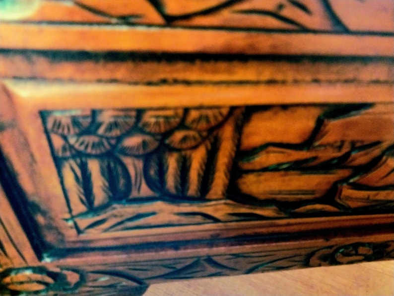 May include: Close-up of a wooden furniture piece with intricate, dark-colored carvings. The wood has a warm, orange-brown hue, and the carvings feature detailed patterns and shapes, creating a textured surface.
