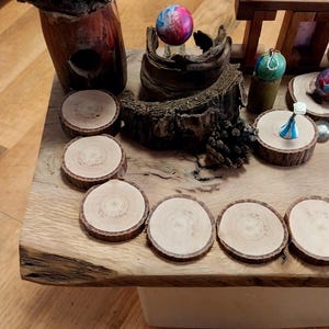 May include: A rustic wooden table displays a collection of handcrafted items. Included are several wooden coasters, a decorative mushroom sculpture, and small, colorful ornaments. The table's natural wood grain and unique items create a warm, inviting aesthetic.