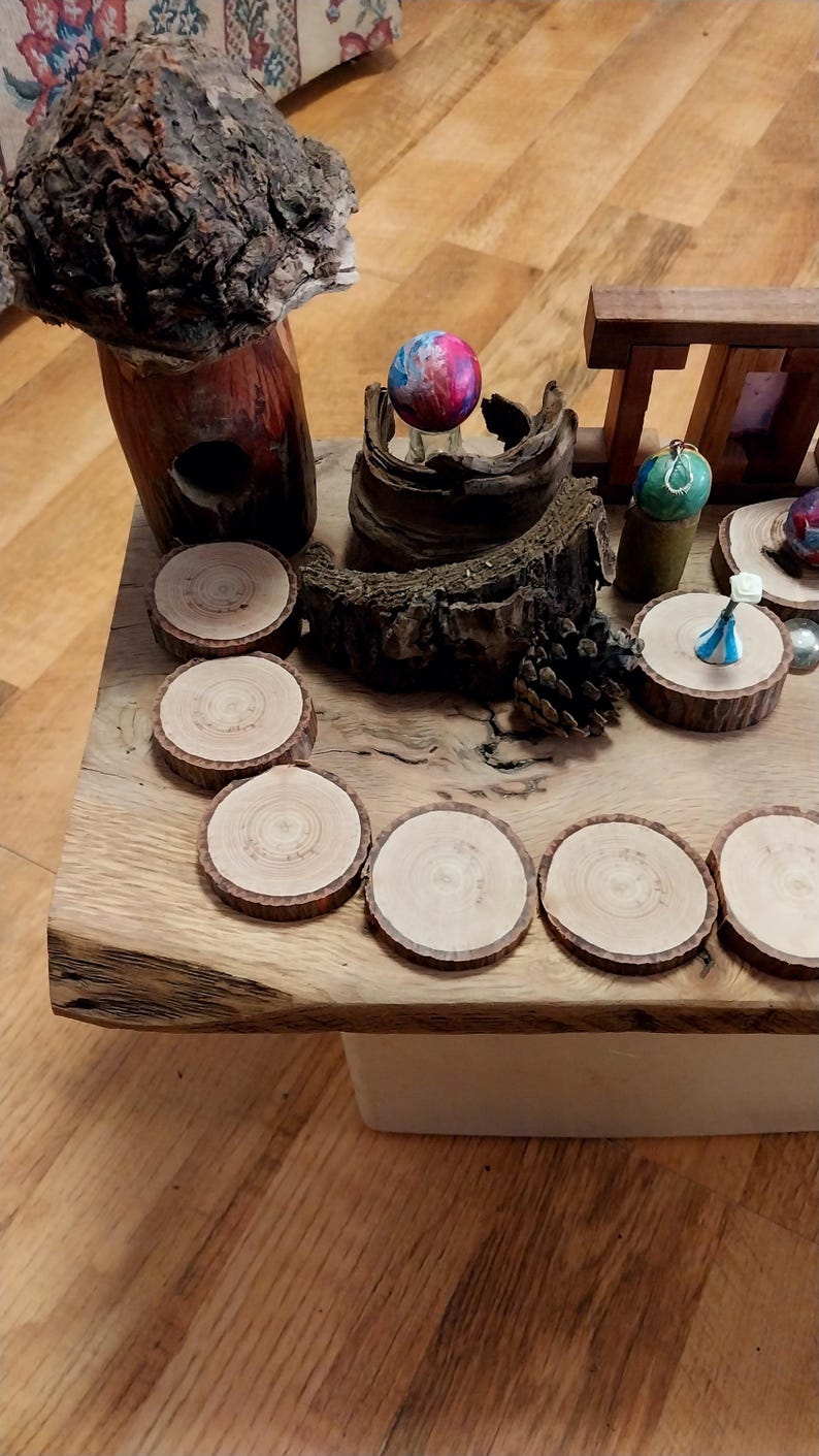 May include: A rustic wooden table displays a collection of handcrafted items. Included are several wooden coasters, a decorative mushroom sculpture, and small, colorful ornaments. The table's natural wood grain and unique items create a warm, inviting aesthetic.