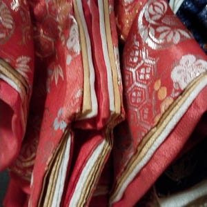 May include: Close-up of a red and gold traditional Japanese garment. The fabric features intricate floral and geometric patterns in silver and white. The garment has layered sleeves and a striped front panel with gold and white accents.