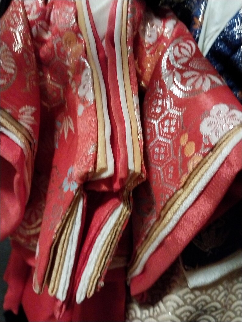 May include: Close-up of a red and gold traditional Japanese garment. The fabric features intricate floral and geometric patterns in silver and white. The garment has layered sleeves and a striped front panel with gold and white accents.