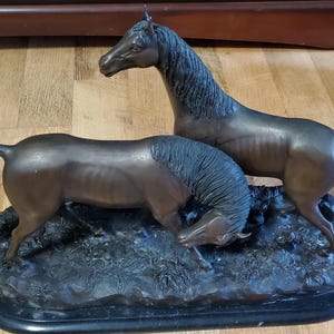 May include: A bronze sculpture of two horses. One horse stands upright, while the other is lowered, appearing to graze. The sculpture is mounted on a black base, with detailed depictions of foliage. The horses are a dark brown color.