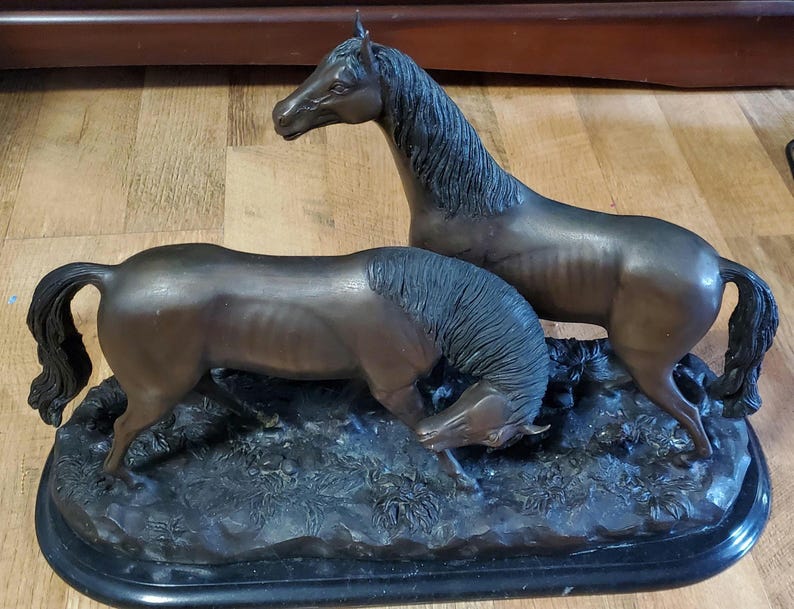 May include: A bronze sculpture of two horses. One horse stands upright, while the other is lowered, appearing to graze. The sculpture is mounted on a black base, with detailed depictions of foliage. The horses are a dark brown color.