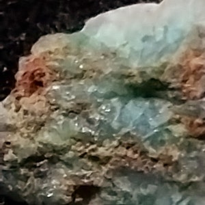 May include: A close-up of a rough, layered rock with a mix of green, brown, and white colors. The rock has a crystalline texture and appears to be a natural mineral specimen.