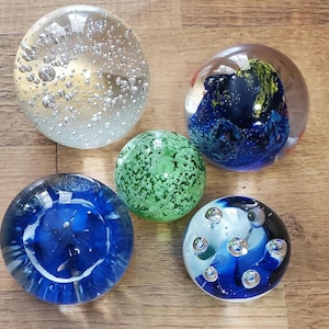 May include: Five glass paperweights of varying designs. One is clear with bubbles, another is green with a speckled pattern, and two are blue with internal designs. The fifth paperweight has a blue and white swirl design with bubbles.
