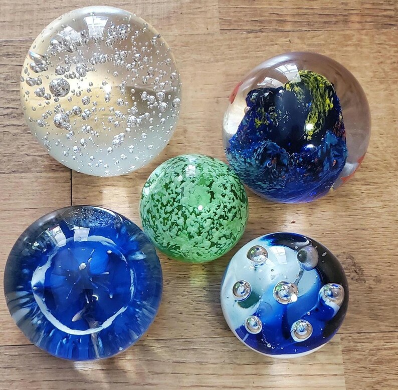 May include: Five glass paperweights in different designs. One is clear with bubbles, another is green with a speckled pattern, and two are blue with internal designs. The fifth has a blue and white swirl design with bubbles.