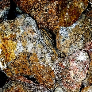 May include: Close-up of a collection of various rocks. The rocks display a mix of colors, including shades of brown, gold, gray, and hints of red. The textures vary, with some appearing smooth and others rough and granular.