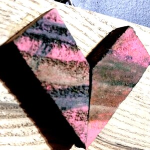 May include: Two triangular pieces of wood with a layered, striped pattern in shades of pink, brown, black, and green. The wood has a rough, textured surface.