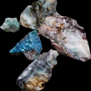 May include: A collection of rough, raw mineral specimens. The stones are a mix of white, blue, green, and brown.