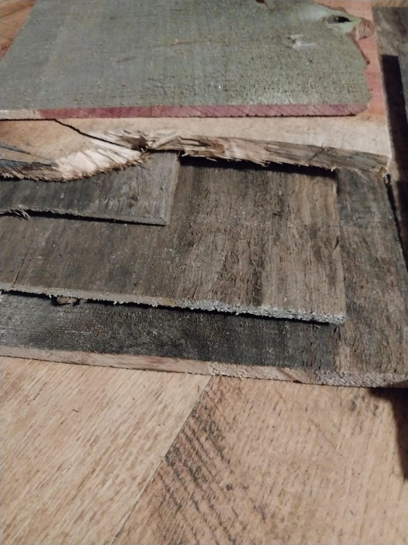 May include: A collection of weathered, reclaimed wood planks in varying sizes and shades of gray and brown. Some planks have visible grain and texture, with rough edges and signs of wear. The wood appears to be repurposed.