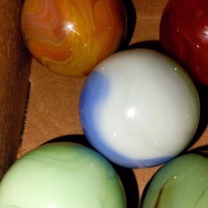 May include: A collection of colorful glass marbles. The marbles are various colors, including orange, red, green, and blue. The marbles are in a brown box. The marbles have a swirled pattern.
