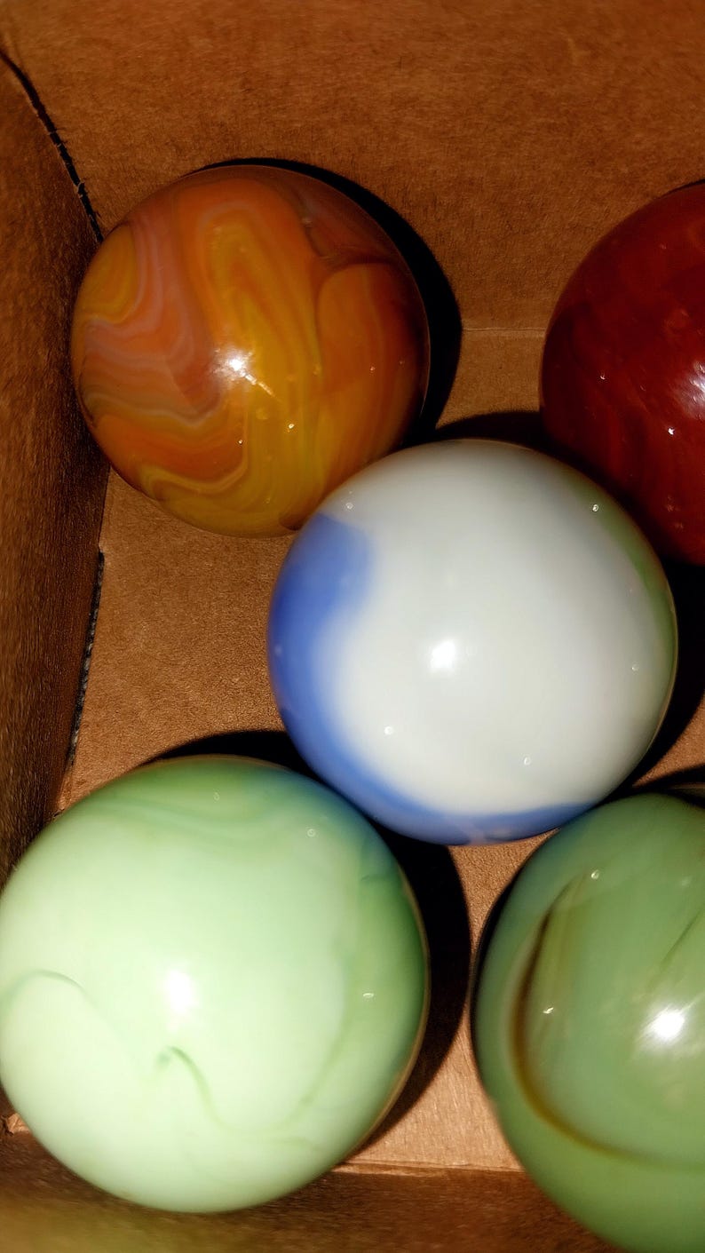 May include: A collection of colorful glass marbles. The marbles are various colors, including orange, red, green, and blue. The marbles are in a brown box. The marbles have a swirled pattern.