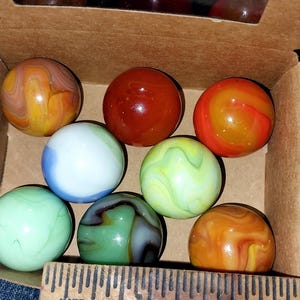 May include: A collection of colorful glass marbles in a cardboard box. The marbles are various colors, including orange, red, green, blue, and white, with swirled patterns. A wooden ruler is visible at the bottom of the box.