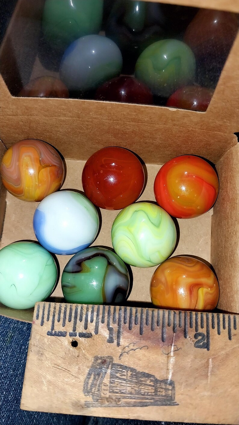 May include: A collection of colorful glass marbles in a cardboard box. The marbles are various colors, including orange, red, green, blue, and white, with swirled patterns. A wooden ruler is visible at the bottom of the box.