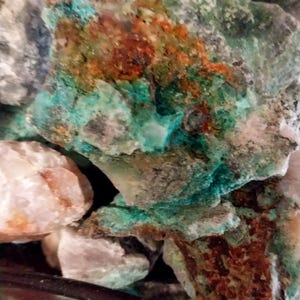 May include: Close-up of a collection of raw, uncut stones. The stones display a variety of colors, including shades of green, brown, and white. The textures vary from rough to smooth, showcasing natural mineral formations.