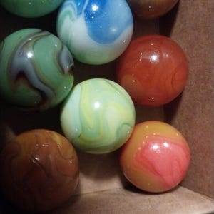 May include: A collection of colorful glass marbles in a cardboard box. The marbles feature swirls of green, blue, orange, and yellow. The marbles are round and have a glossy finish. The box has a ruler printed on the side.