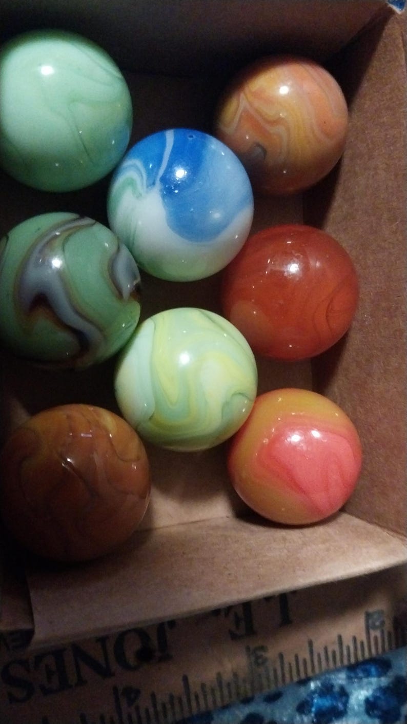 May include: A collection of colorful glass marbles in a cardboard box. The marbles feature swirls of green, blue, orange, and yellow. The marbles are round and have a glossy finish. The box has a ruler printed on the side.