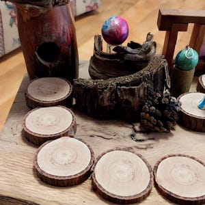 May include: A collection of natural wood coasters with a dark brown bark edge. The coasters are arranged on a rustic wooden surface, alongside decorative wooden sculptures and a colorful, spherical ornament. A pine cone adds to the natural theme.