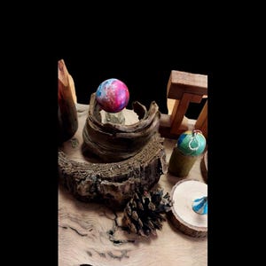 May include: A collection of decorative spheres and wooden elements. A colorful sphere with pink, blue, and purple hues sits atop a wooden base. Other spheres with green and blue accents are displayed on wooden stands. The scene is set on a wooden surface.