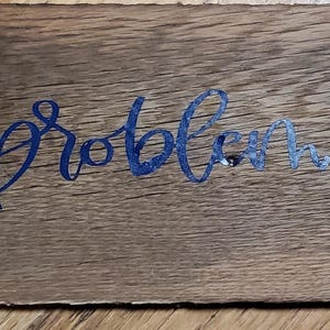 May include: A wooden sign displaying the text "I'm the problem, it's me" in blue and silver script. The sign is made from aged wood, showing the grain and small holes, giving it a rustic appearance.