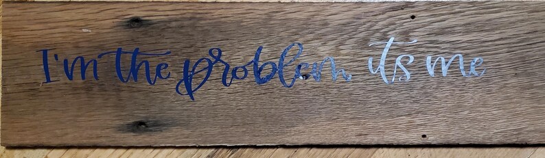 May include: A wooden sign displaying the text "I'm the problem, it's me" in blue and silver script. The sign is made from aged wood, showing the grain and small holes, giving it a rustic appearance.